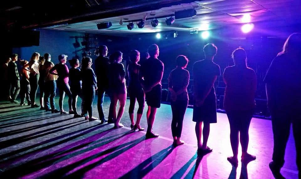 The backs of 15 performers on the stage at Chico Theater Company, who are illuminated by purple and blue lights and stare out at the audience