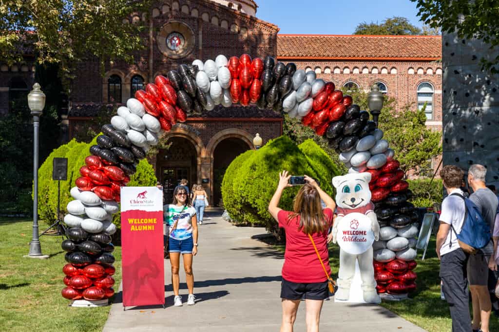 A woman poses under a balloon arch in front of a "Welcome Alumni" sign while another woman takes her picture during Chico State's Wildcat Weekend alumni event