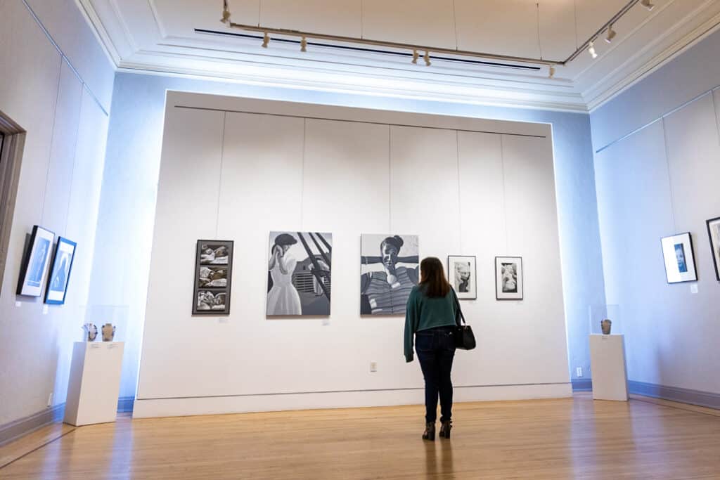 A woman stands in the middle of the Museum of Northern California Art (monca) looking at hanging portraits