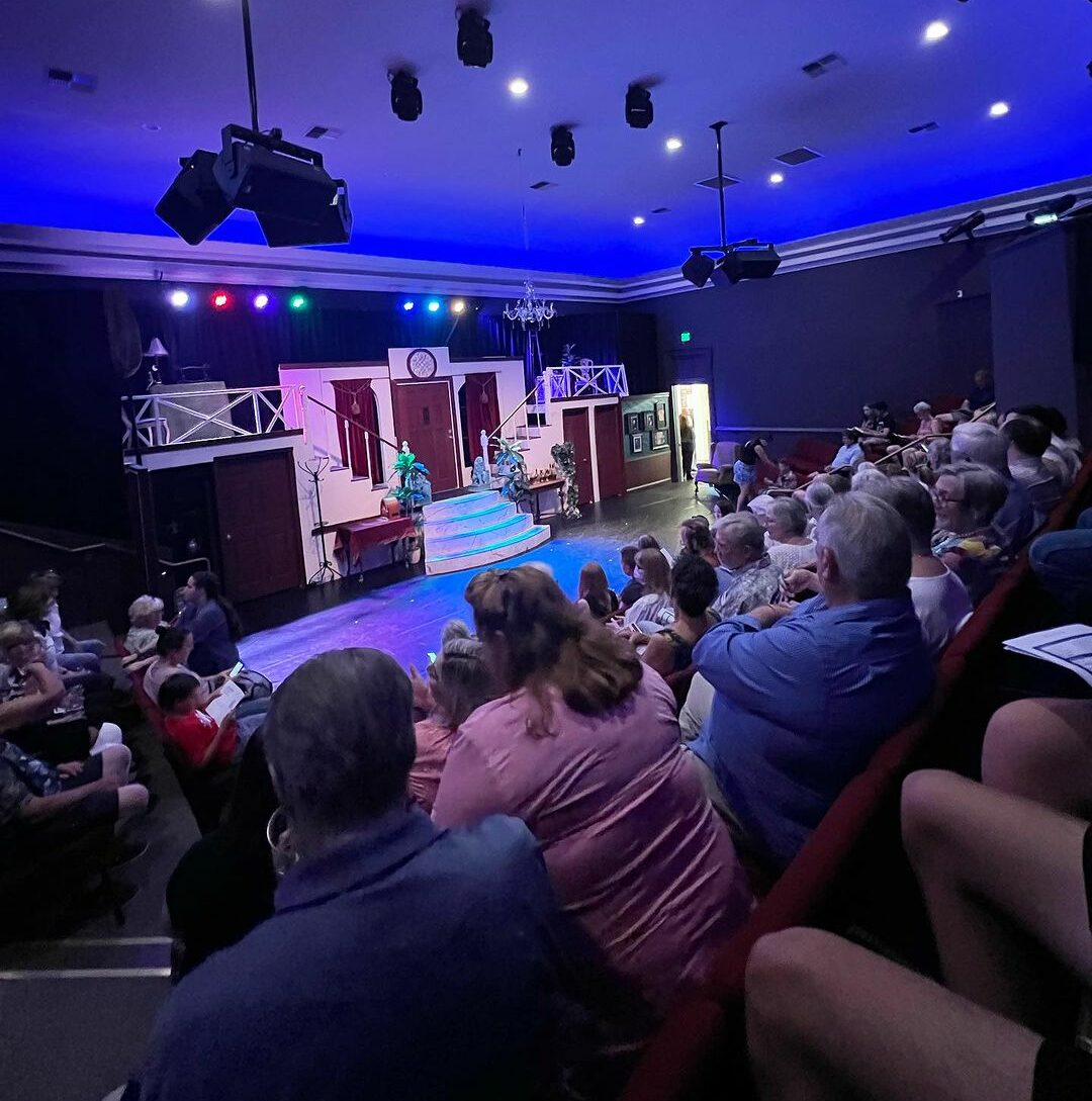 A view of a small, intimate theater stage from the audience, with rows of audience members in the foreground