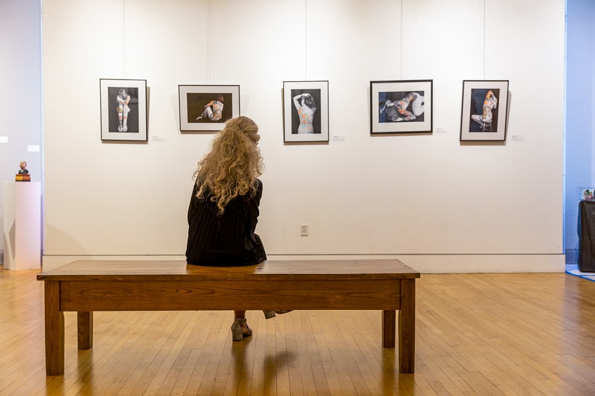 A woman sits on a bench looking at a gallery wall with five illustrations at the Museum of Northern California Art