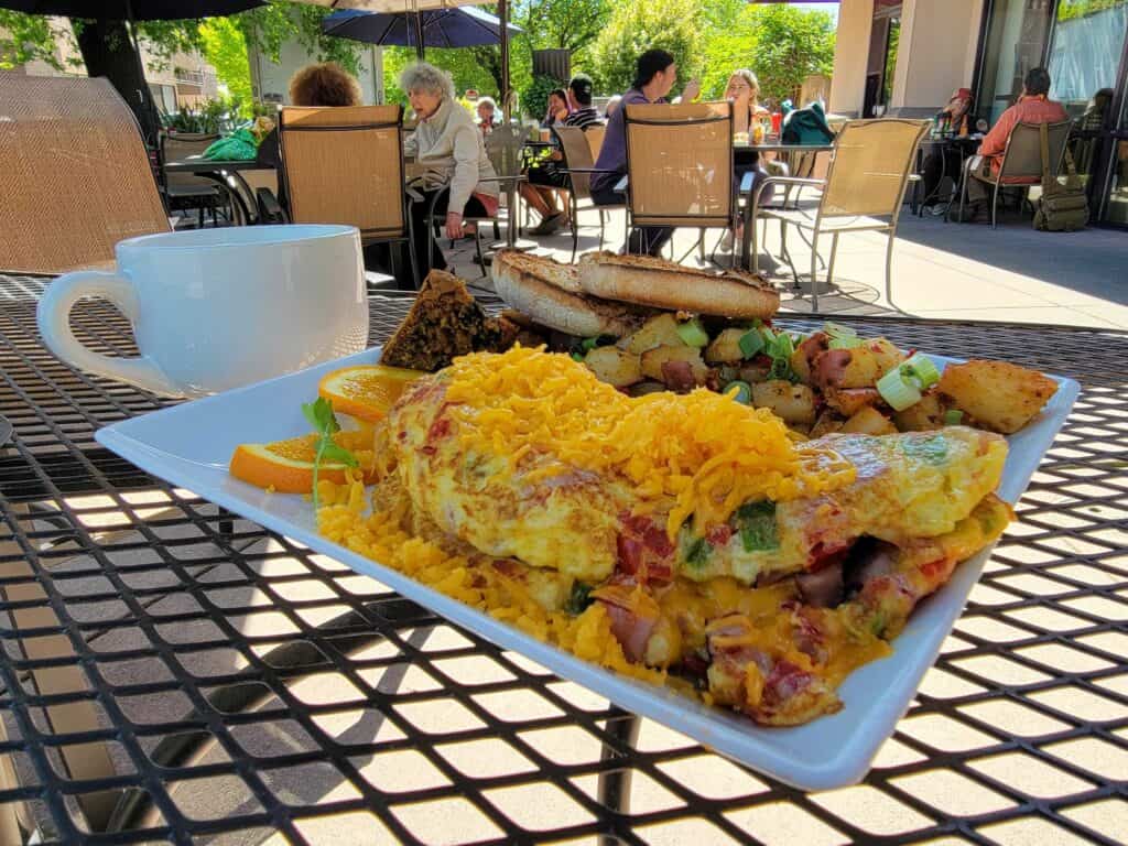 A closeup of a loaded omelette with country potatoes on an outside table with diners in the background at Beatniks Coffee House & Breakfast Joint