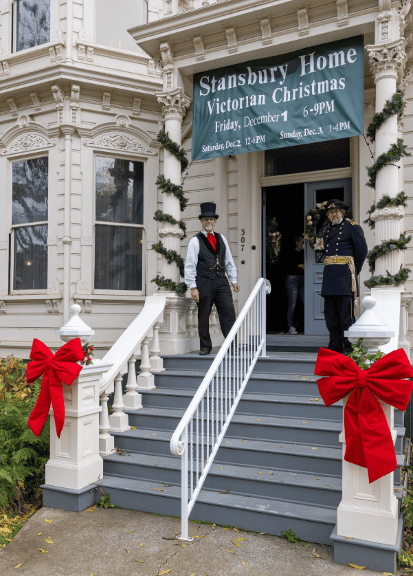 The Stansbury Home Victorian Christmas 7 Two older white men in formal Victorian era suits flank the front doors of the Stansbury home, a large white Victorian mansion decorated with green garlands on pillars, red bows on the entrance handrail posts, and a vinyl sign above the door with details about the Stansbury Home Victorian Christmas event.