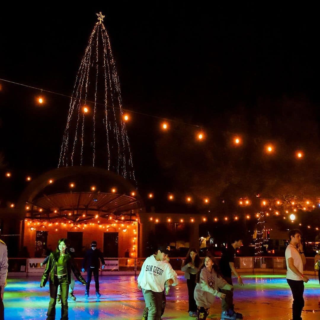Downtown Chico Community Christmas Tree Lighting 7 On the night of the Downtown Chico Community Christmas Tree Lighting, young adults ice skate in the foreground wearing stylish winter clothing at night, lit up with colorful lights and a tall tree stands in the background decorated with white Christmas lights and a star.